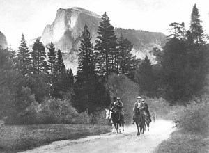 President Theodore Roosevelt and John Muir riding horses along a road in Yosemite Valley, with Half Dome in the distance, accompanied by Park Rangers Archie Leonard and Charles Leidig, followed by unidentified man on foot; left to right, Leonard, Muir, Roosevelt, Leidig.